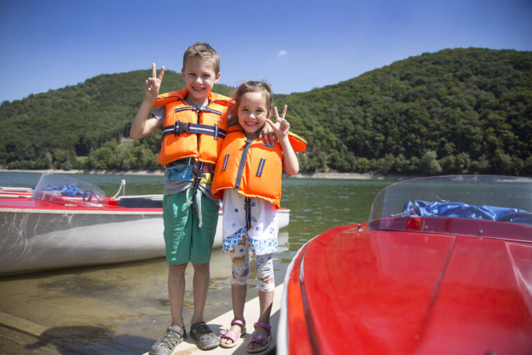 Kinder in bunten Kleidern tragen Rettungswesten und stehen vor einem rot-weißen Boote am Diemelsee. Sie befinden sich auf dem Bootssteg, und der strahlend blaue Himmel lässt die sommerliche Atmosphäre perfekt zur Geltung kommen. Die Kinder freuen sich auf ihren Ausflug auf dem Wasser, umgeben von der idyllischen Natur des Diemelsees.