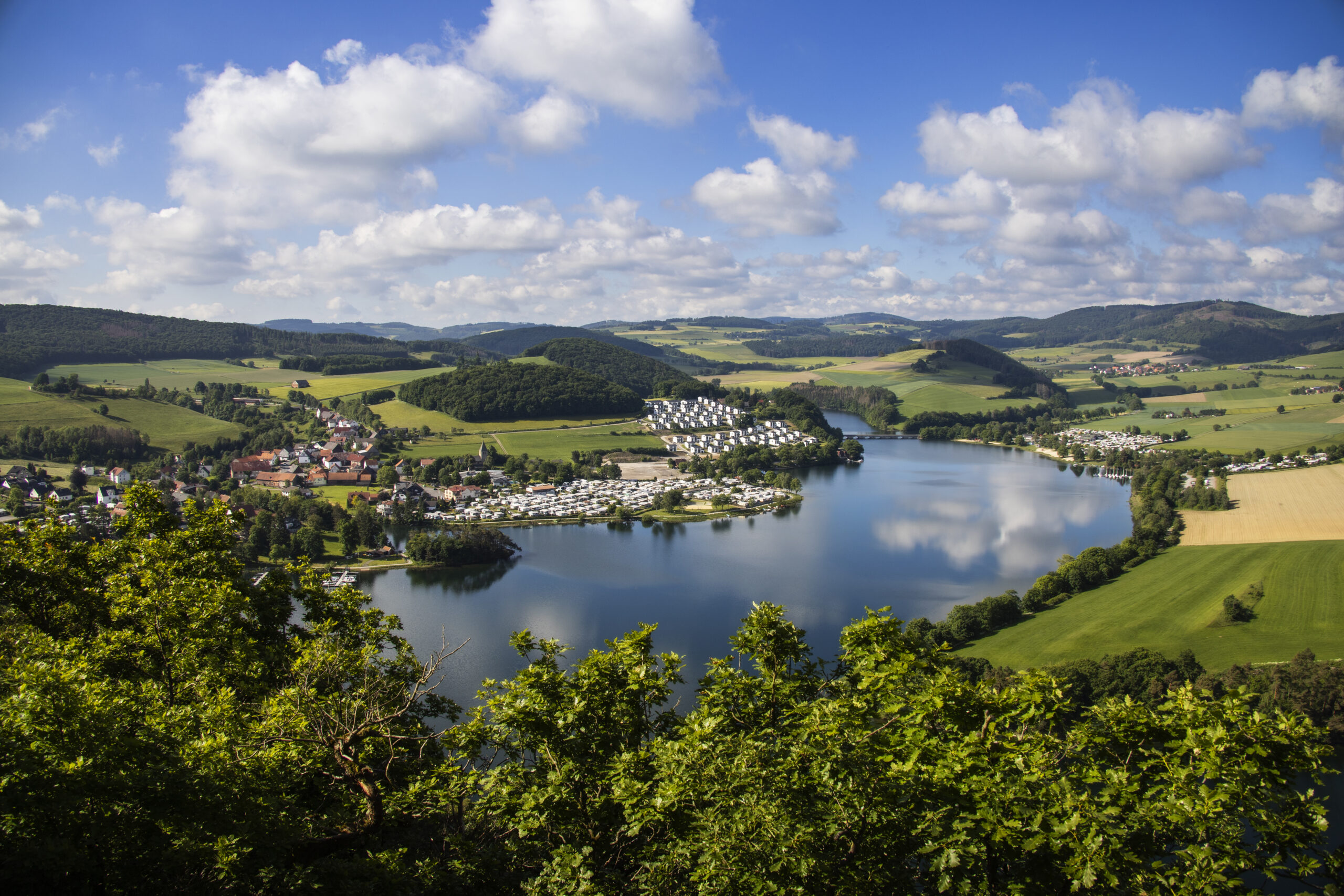 Blick vom Aussichtspunkt St. Muffert auf den Diemelsee. Der Blick vom Aussichtspunkt St. Muffert zeigt den weitläufigen Diemelsee inmitten einer grünen Landschaft. Der See glitzert im Sonnenlicht, während sich sanfte Hügel und Wälder rundherum erstrecken. Der Himmel ist mit Wolken bedeckt, und die Umgebung wirkt friedlich und naturverbunden.