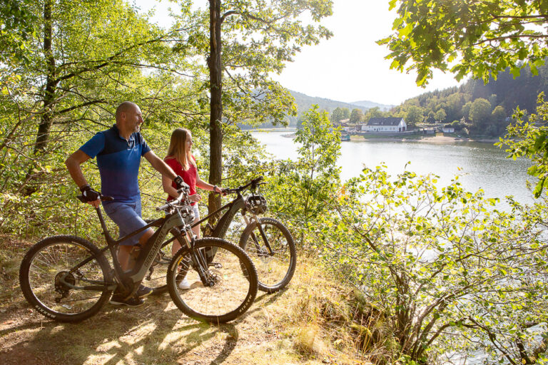 Zwei Radfahrer stehen mit ihren E-Bikes in einem Waldgebiet am Diemelsee. Zwischen grünen Bäumen und Sträuchern blicken sie auf den schimmernde Diemelsee. Die Radfahrer tragen auffällige, bunte Kleidung – einer in Blau, der andere in Rot – und genießen die frische Luft und die Aussicht. Die Szene strahlt eine ruhige, aktive Atmosphäre aus, ideal für Natur- und Radfahrbegeisterte.