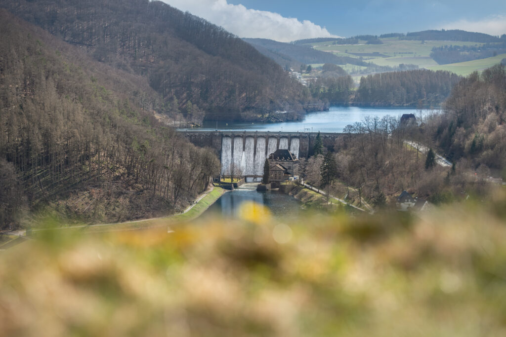 Das Bild zeigt den Diemelsee mit seiner imposanten Staumauer im Vordergrund. Wasser fließt kraftvoll über den Überlauf der Staumauer und erzeugt spritzende Gischt. Die Umgebung ist geprägt von grünen Wäldern und sanften Hügeln. Der See selbst spiegelt den Himmel und vermittelt eine ruhige, natürliche Atmosphäre. Die technische Konstruktion der Staumauer verbindet sich harmonisch mit der umliegenden Landschaft und zeigt die Bedeutung des Diemelsees als Wasserspeicher und Naherholungsgebiet.