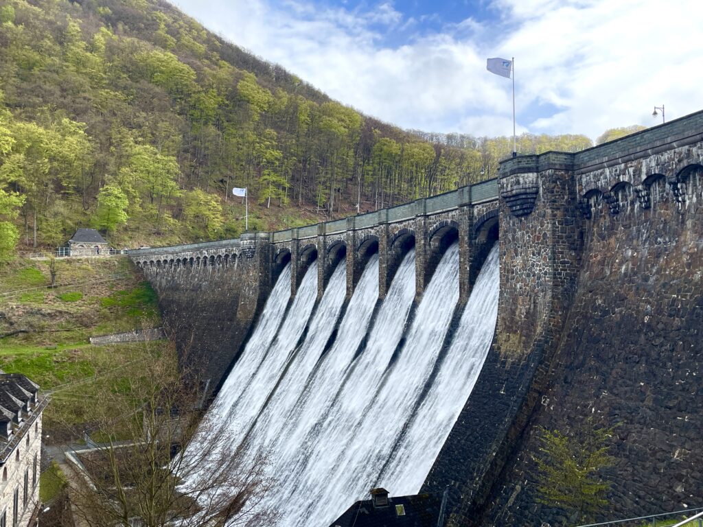 Das Bild zeigt den Überlauf der Diemeltalsperre in der Jahreszeit Frühjahr. Aufgrund der Schneeschmelze und vermehrten Niederschläge fließt das Wasser kraftvoll und in großer Menge über die Staumauer. Die Umgebung ist geprägt von frischem, grünem Laub und jungen Pflanzen. Die spritzende Gischt und das bewegte Wasser vermitteln die lebendige Kraft des Wasserkreislaufs und die Bedeutung der Talsperre als Wasserspeicher und Schutzanlage in der Region.