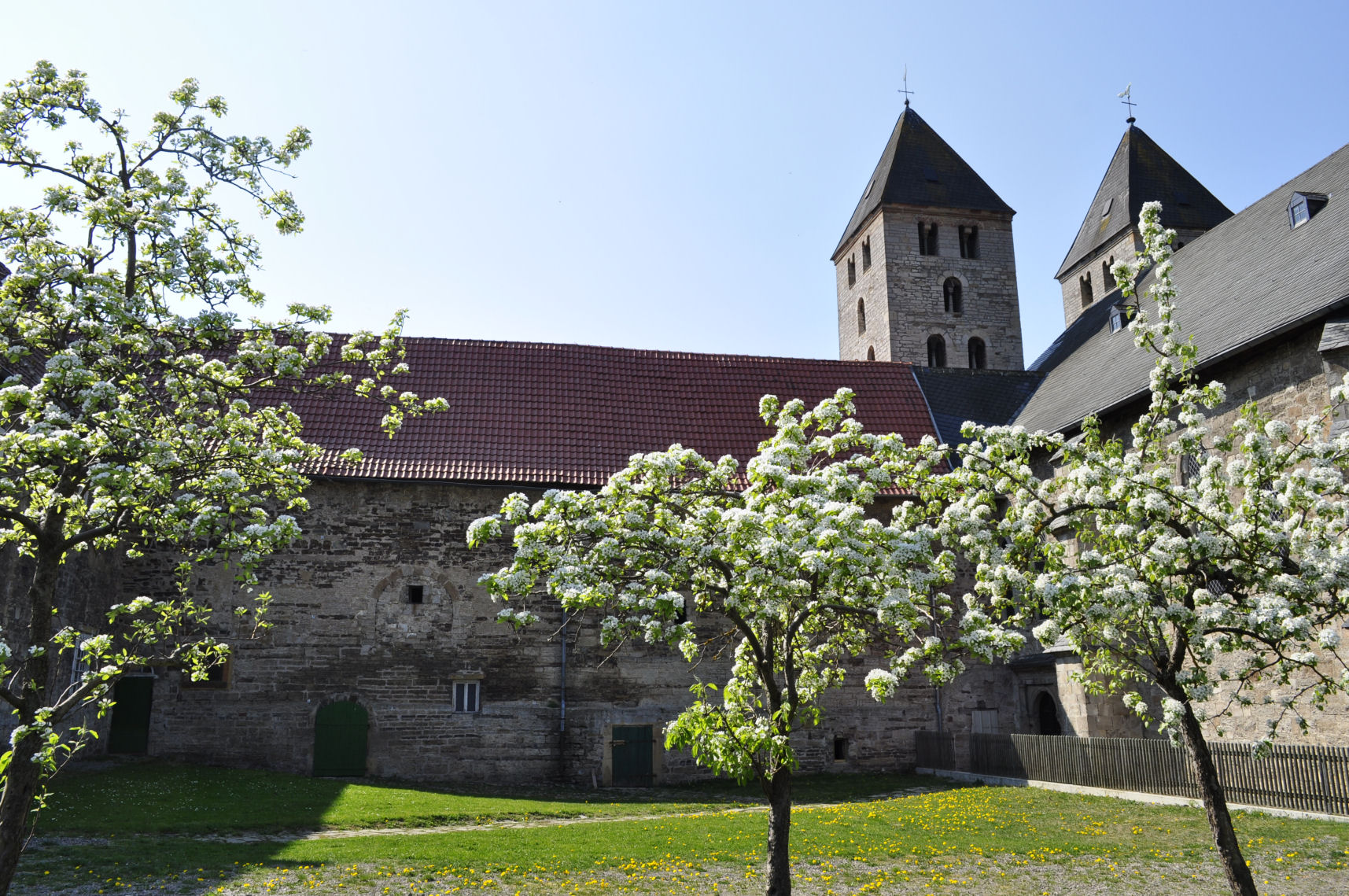 Das Bild zeigt das Kloster Flechtdorf in der Frühlingszeit. Die hohen Türme des historischen Gebäudes ragen stolz in den hellen Himmel. Vor dem Kloster blühen Kirchbäume, deren weißen Blüten die Szenerie lebendig und freundlich wirken lassen. Die frische Jahreszeit ist durch das zarte Grün der Blätter und die Blütenpracht deutlich spürbar. Die Atmosphäre lädt zu einem Ausflug und zur Besichtigung des Klosters ein.