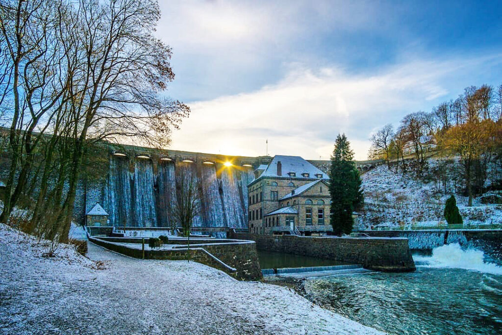 Das Bild zeigt den Überlauf der Diemeltalsperre im Winter. Wasser fließt über die Staumauer, während sich an den Rändern Eis bildet und die umliegende Landschaft mit Schnee bedeckt ist. Die kalte Jahreszeit verleiht der Szene eine ruhige, fast stille Atmosphäre, obwohl das Wasser unaufhörlich weiterfließt. Die Kombination aus Wasser, Eis und Schnee zeigt den Jahreszeitenwechsel und die Funktion der Talsperre auch bei kalten Temperaturen.