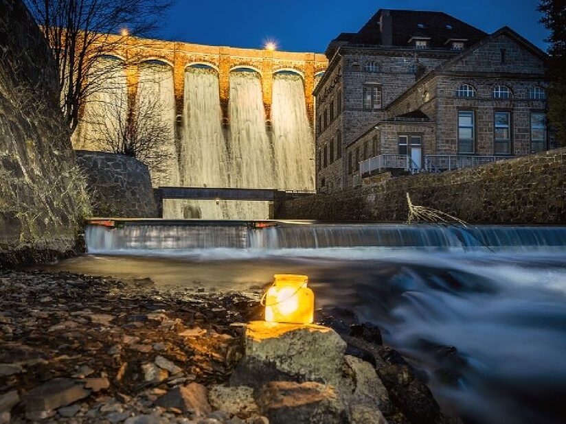 Das Bild zeigt die imposante Staumauer des Diemelsees in nächtlicher Stimmung. Die Mauer ist von mehreren gelblich leuchtenden Lampen erhellt, die ihre Struktur betonen. Im Vordergrund steht eine klassische Laterne, die ebenfalls Licht ausstrahlt und dem Bild eine stimmungsvolle Tiefe verleiht. Vor der Mauer ist das Kraftwerks zu erkennen. Die Szene verbindet Technik und Atmosphäre und verleiht der Staumauer eine fast mystische Ausstrahlung in der Dunkelheit.