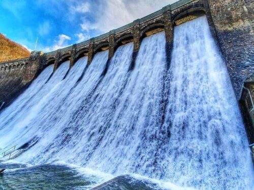 Das Bild zeigt den Überlauf der Staumauer am Diemelsee an einem sonnigen Tag. Große Wassermengen strömen kraftvoll über den Rand der Staumauer und erzeugen eine spritzende Gischt. Die Struktur der Staumauer ist gut sichtbar, und die umgebende Landschaft wirkt hell und klar. Die Szene vermittelt die beeindruckende Kraft des Wassers und die technische Bedeutung des Staubeckens für die Region.