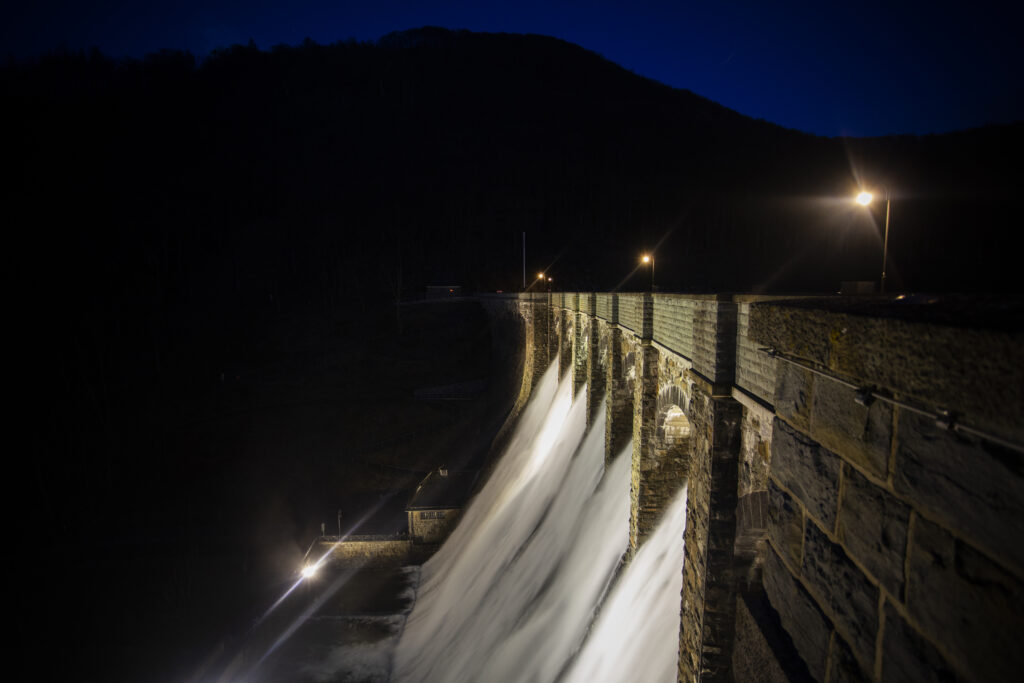 Das Bild zeigt die Staumauer des Diemelsees bei Nacht. Über den Überlauf der Mauer strömt Wasser, das im Schein der Lampen sanft beleuchtet wird. Die Lichter entlang der Staumauer und der angrenzenden Wege verleihen der Szene eine ruhige, fast mystische Atmosphäre. Der dunkle Himmel und die reflektierenden Wassermassen erzeugen einen starken Kontrast, der die technische Konstruktion und die natürliche Umgebung eindrucksvoll in Szene setzt.