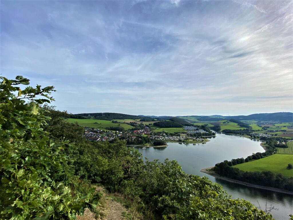 Das Bild zeigt einen weiten Blick über den Diemelsee an einem sonnigen Sommertag. Der Himmel ist klar und blau. Das Wasser des Sees glitzert in der Sonne. Rund um den See erstrecken sich grüne Felder, Wiesen und Bäume. Im Hintergrund liegt der Ort Heringhausen mit mehreren hellen Häusern. Eine Brücke überspannt den See und verbindet die Ufer. Die Landschaft wirkt ruhig und idyllisch – eine typische Sommerstimmung im Sauerland.