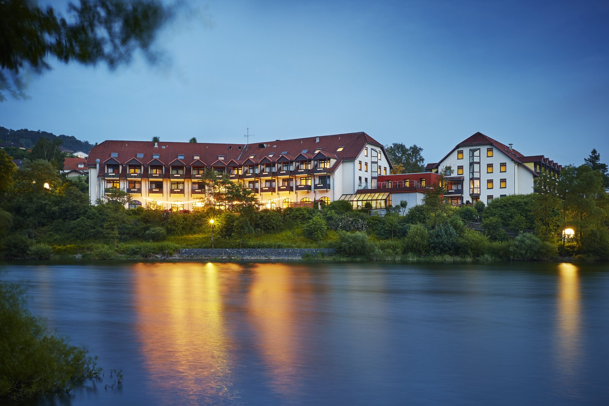 Das Bild zeigt das Hotel Göbel am Diemelsee im Sauerland bei Nacht. Das Hotel liegt direkt am Seeufer, umgeben von grüner Vegetation. Die Außenbeleuchtung des Hotels spiegelt sich sanft im ruhigen Wasser des Sees und schafft eine einladende und atmosphärische Stimmung.