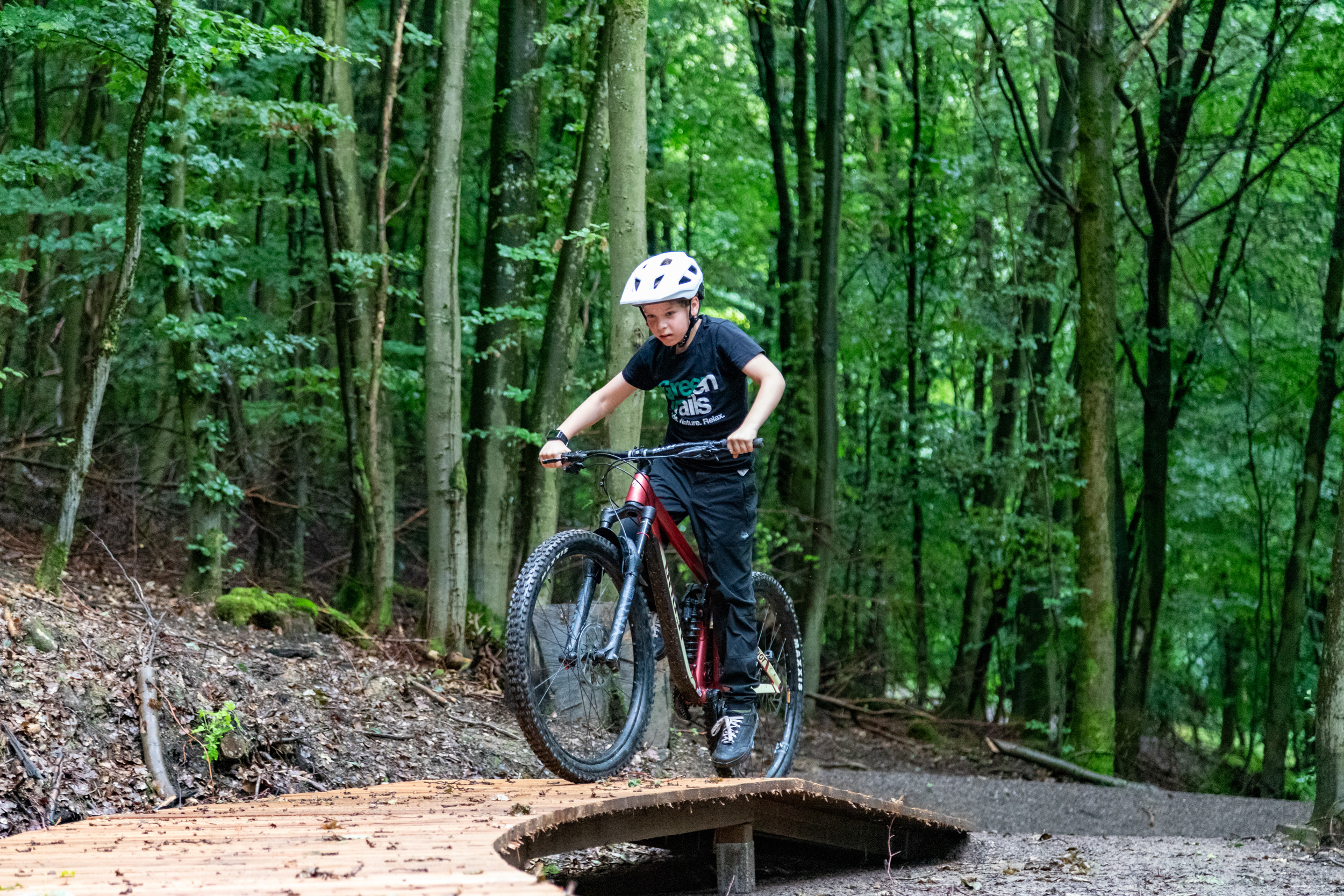 Das Bild zeigt ein Kind, das sportlich in dunkler Kleidung und mit Helm auf einem roten Fahrrad die Green Trails im Wald fährt. Es fährt über ein Podest, das den Blick auf den Wald freigibt. Die Umgebung ist grün und naturbelassen, und das Kind wirkt konzentriert und bereit für die nächste Etappe auf der Trail-Strecke. Das Bild zeigt ein Kind, das sportlich in dunkler Kleidung und mit Helm auf einem roten Fahrrad die Green Trails im Wald fährt. Es fährt über ein Podest, das den Blick auf den Wald freigibt. Die Umgebung ist grün und naturbelassen, und das Kind wirkt konzentriert und bereit für die nächste Etappe auf der Trail-Strecke.