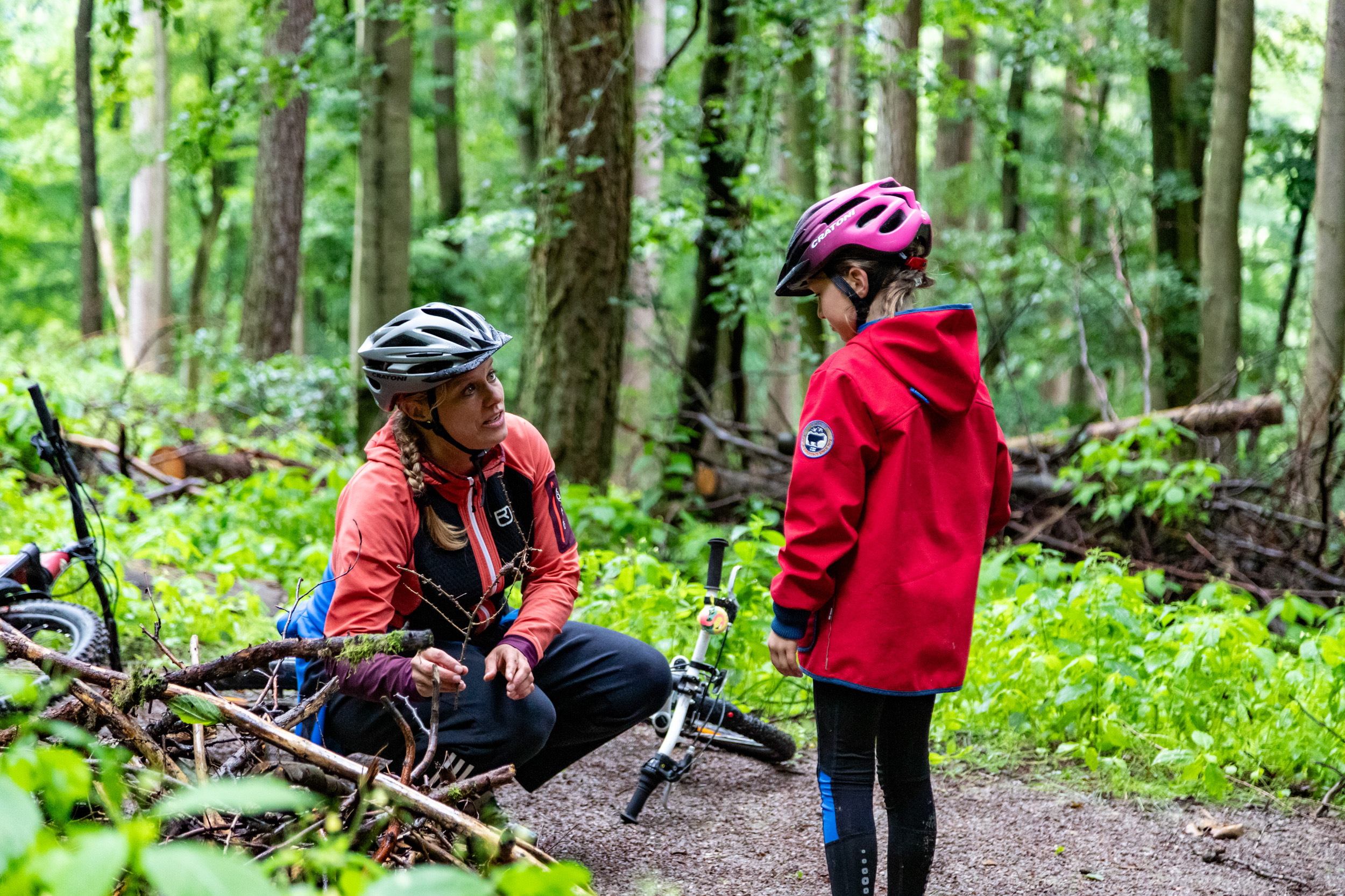 Das Bild zeigt eine Mutter und ihre Tochter auf dem Biberpfad, einem Themenweg der Green Trails am Diemelsee. Beide tragen rote Jacken und dunkle Hosen. Ihre Fahrräder liegen am Rand des naturbelassenen Wegs. Die Mutter zeigt der Tochter etwas am Wegesrand und erklärt es ihr – vielleicht eine Spur des Bibers. Die Szene spielt sich in einer waldreichen Umgebung ab und vermittelt eine ruhige, lehrreiche Familienatmosphäre beim Radfahren in der Natur. Das Bild zeigt eine Mutter und ihre Tochter auf dem Biberpfad, einem Themenweg der Green Trails am Diemelsee. Beide tragen rote Jacken und dunkle Hosen. Ihre Fahrräder liegen am Rand des naturbelassenen Wegs. Die Mutter zeigt der Tochter etwas am Wegesrand und erklärt es ihr – vielleicht eine Spur des Bibers. Die Szene spielt sich in einer waldreichen Umgebung ab und vermittelt eine ruhige, lehrreiche Familienatmosphäre beim Radfahren in der Natur.