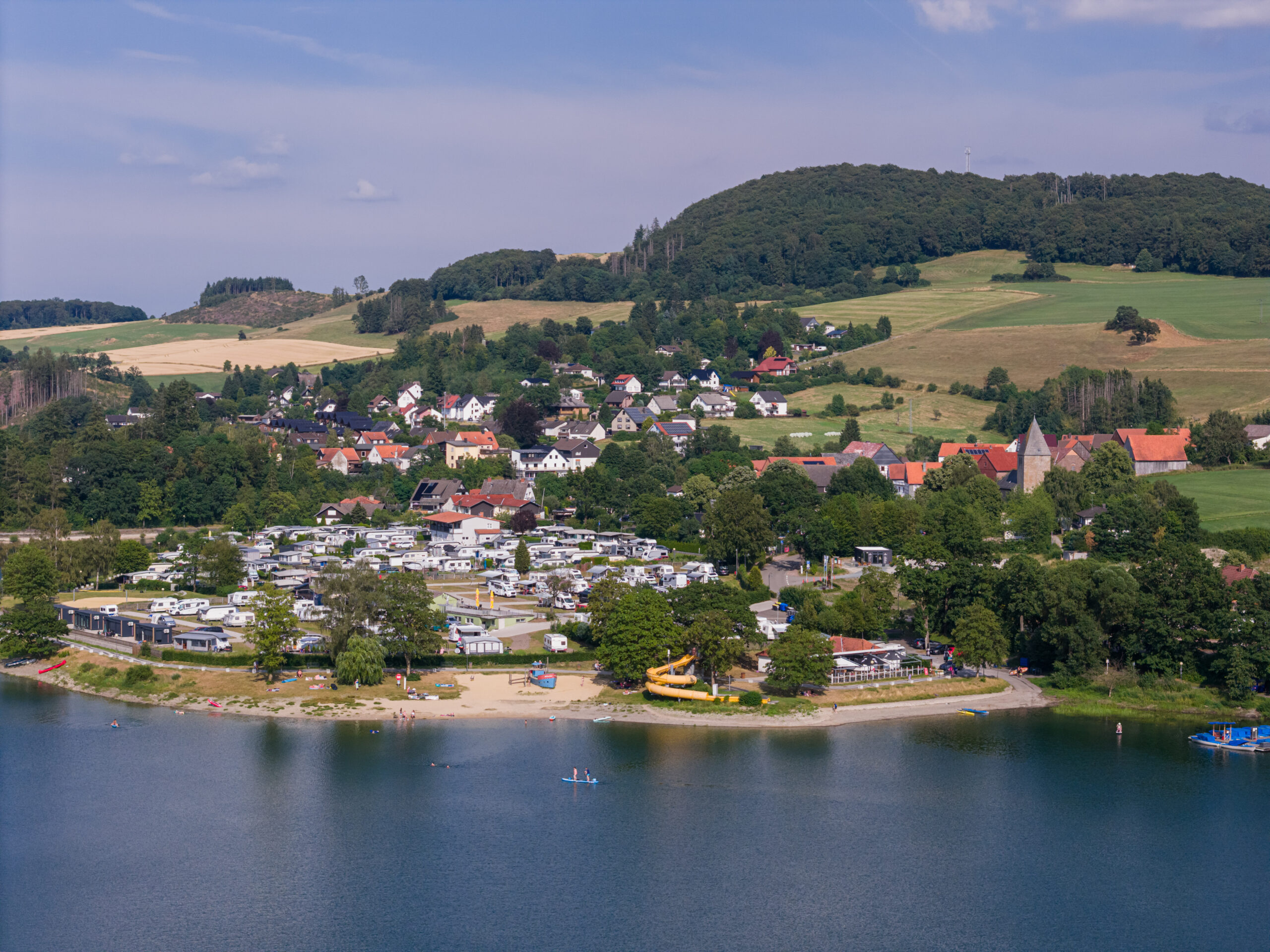 Eine Luftaufnahme zeigt den Bereich Goldbreite am Diemelsee. Im Fokus steht der Campingplatz mit zahlreichen Wohnwagen, umgeben von viel Grün. Im Hintergrund ist der See sichtbar, der einen Seeblick bietet. Die Szene vermittelt eine friedliche Atmosphäre mit Natur und Freizeitaktivitäten am Wasser.