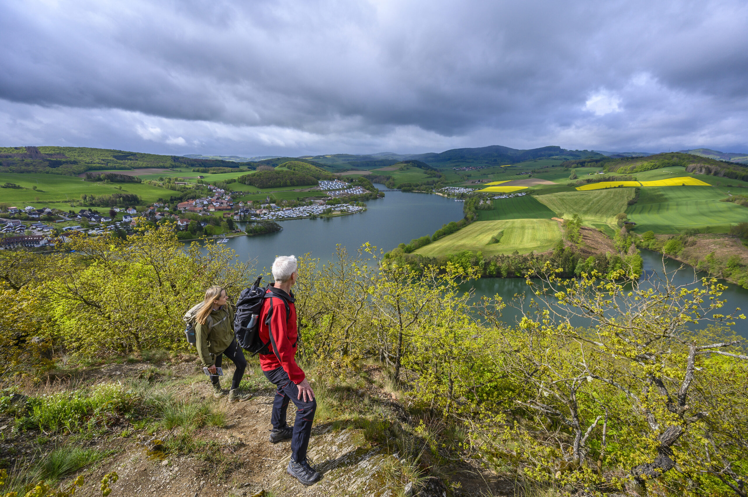 Das Bild zeigt zwei Wanderer, die auf einem Pfad zum Gipfel unterwegs sind. Von dort aus eröffnet sich ein beeindruckender Panoramablick auf das Dorf Heringhausen, den Diemelsee und die umliegenden grünen Wiesen und Wälder. Die Szene strahlt Ruhe und Naturverbundenheit aus und lädt zum Entdecken und Verweilen ein.