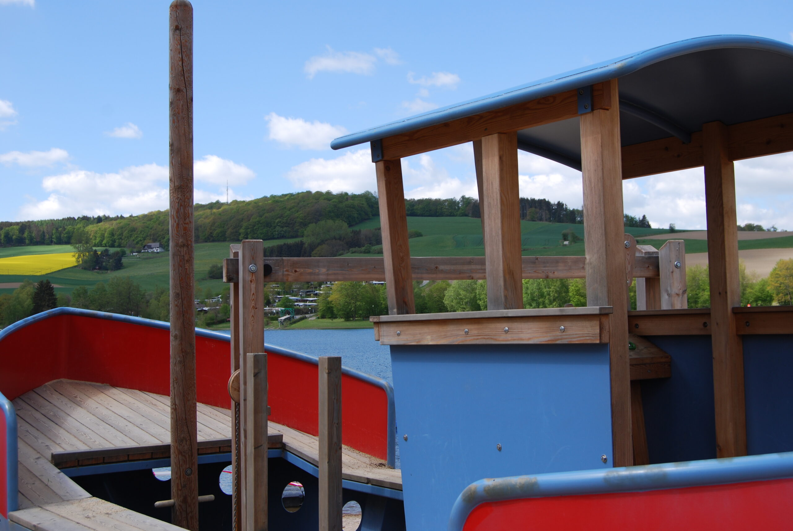 Ein blau-rotes Spielschiff steht am Strandbad in Heringhausen auf dem Sand. Das Spielgerät ist kindgerecht gestaltet und lädt zum Klettern und Spielen ein. Im Hintergrund ist teilweise Wasser und ein Campingplatz zu sehen, das Strandbad wirkt sonnig und einladend. Ein blau-rotes Spielschiff steht am Strandbad in Heringhausen auf dem Sand. Das Spielgerät ist kindgerecht gestaltet und lädt zum Klettern und Spielen ein. Im Hintergrund ist teilweise Wasser und ein Campingplatz zu sehen, das Strandbad wirkt sonnig und einladend.