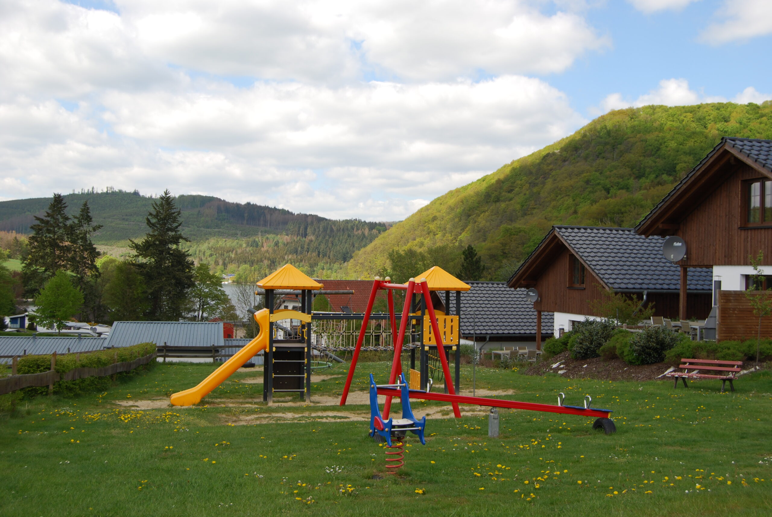 Ein Spielplatz in Heringhausen mit einem Spielturm und einer Wippe. Im Hintergrund öffnet sich der Blick auf den Diemelsee. Rechts sind Häuser zu sehen, umgeben von grünen Wäldern. Der Spielplatz wirkt einladend und naturnah gelegen. Ein Spielplatz in Heringhausen mit einem Spielturm und einer Wippe. Im Hintergrund öffnet sich der Blick auf den Diemelsee. Rechts sind Häuser zu sehen, umgeben von grünen Wäldern. Der Spielplatz wirkt einladend und naturnah gelegen.