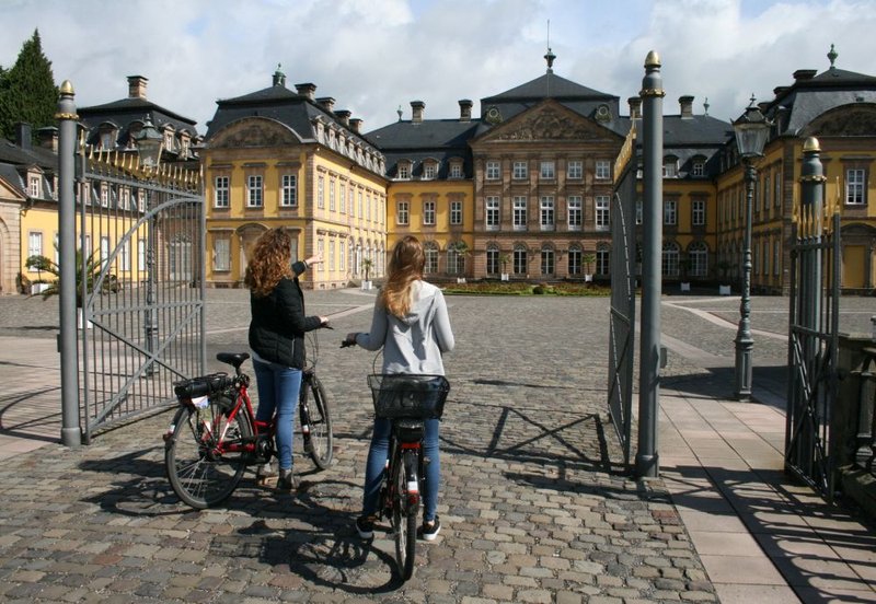 Das Bild zeigt zwei Radfahrerinnen, die vor dem beeindruckenden Rezidenzschloss in Bad Arolsen stehen. Beide sind in gemütlich gekleidet und sind vorn ihren Rädern abgestiegen. Das Schloss im Hintergrund ist ein historisches Gebäude mit barocker Architektur und schönen Gärten, die das Bild umrahmen. Die Radfahrerinnen scheinen eine Pause auf ihrer Radtour zu machen und genießen die Aussicht auf das Schloss.
