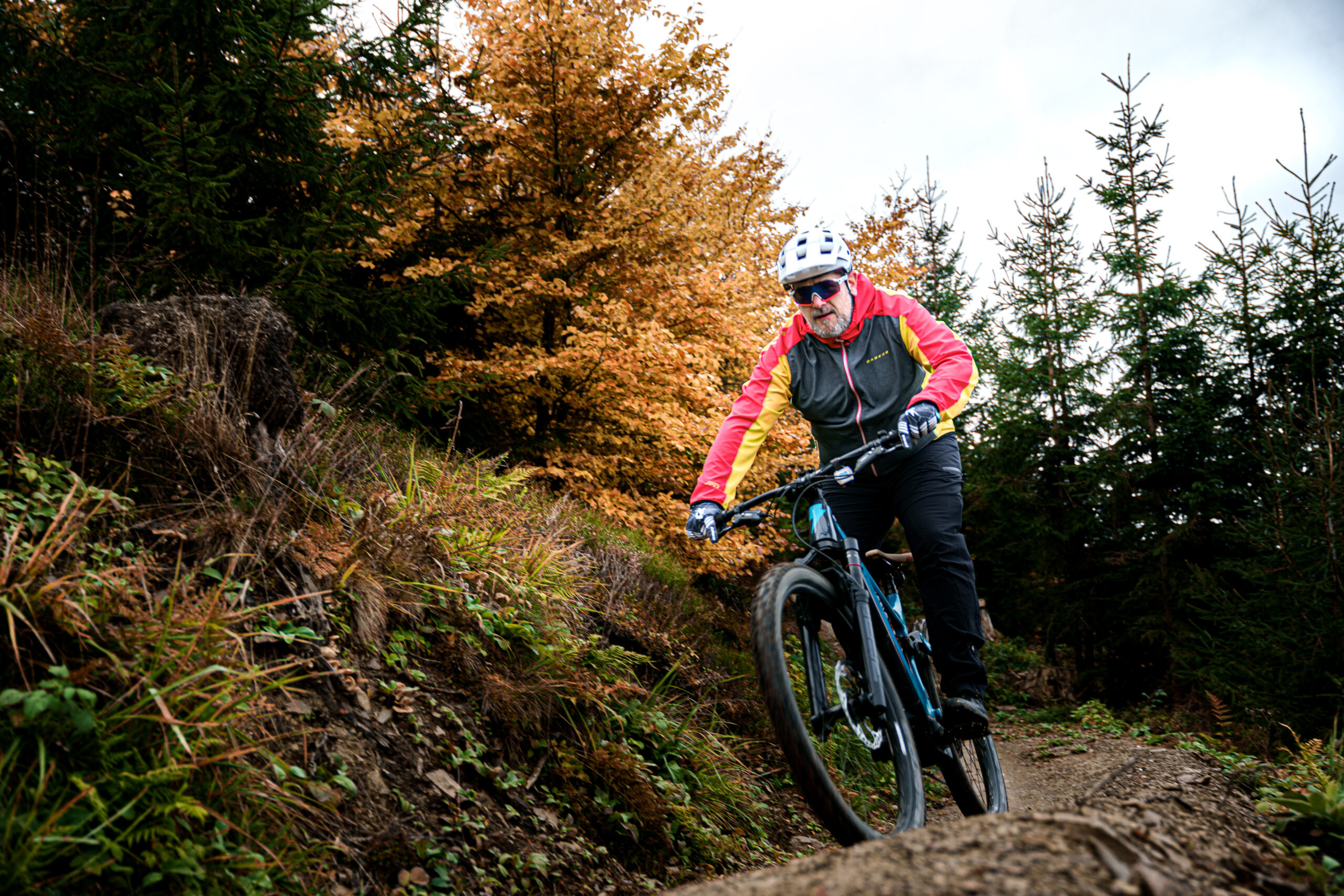 Ein Mountainbiker trägt eine auffällige gelb-rote Jacke, einen Fahrradhelm und eine Sonnenbrille. Er fährt aus einem dichten, schattigen Waldstück heraus auf einem schmalen, befestigten Trail, der leicht bergab verläuft. Der Untergrund ist trocken durchzogen. Im Hintergrund sind hohe Bäume und grünes Buschwerk sichtbar. Der Fahrer ist in Vorwärtsposition, wirkt konzentriert und bewegt sich mit Tempo und Kontrolle. Ein Mountainbiker trägt eine auffällige gelb-rote Jacke, einen Fahrradhelm und eine Sonnenbrille. Er fährt aus einem dichten, schattigen Waldstück heraus auf einem schmalen, befestigten Trail, der leicht bergab verläuft. Der Untergrund ist trocken durchzogen. Im Hintergrund sind hohe Bäume und grünes Buschwerk sichtbar. Der Fahrer ist in Vorwärtsposition, wirkt konzentriert und bewegt sich mit Tempo und Kontrolle.