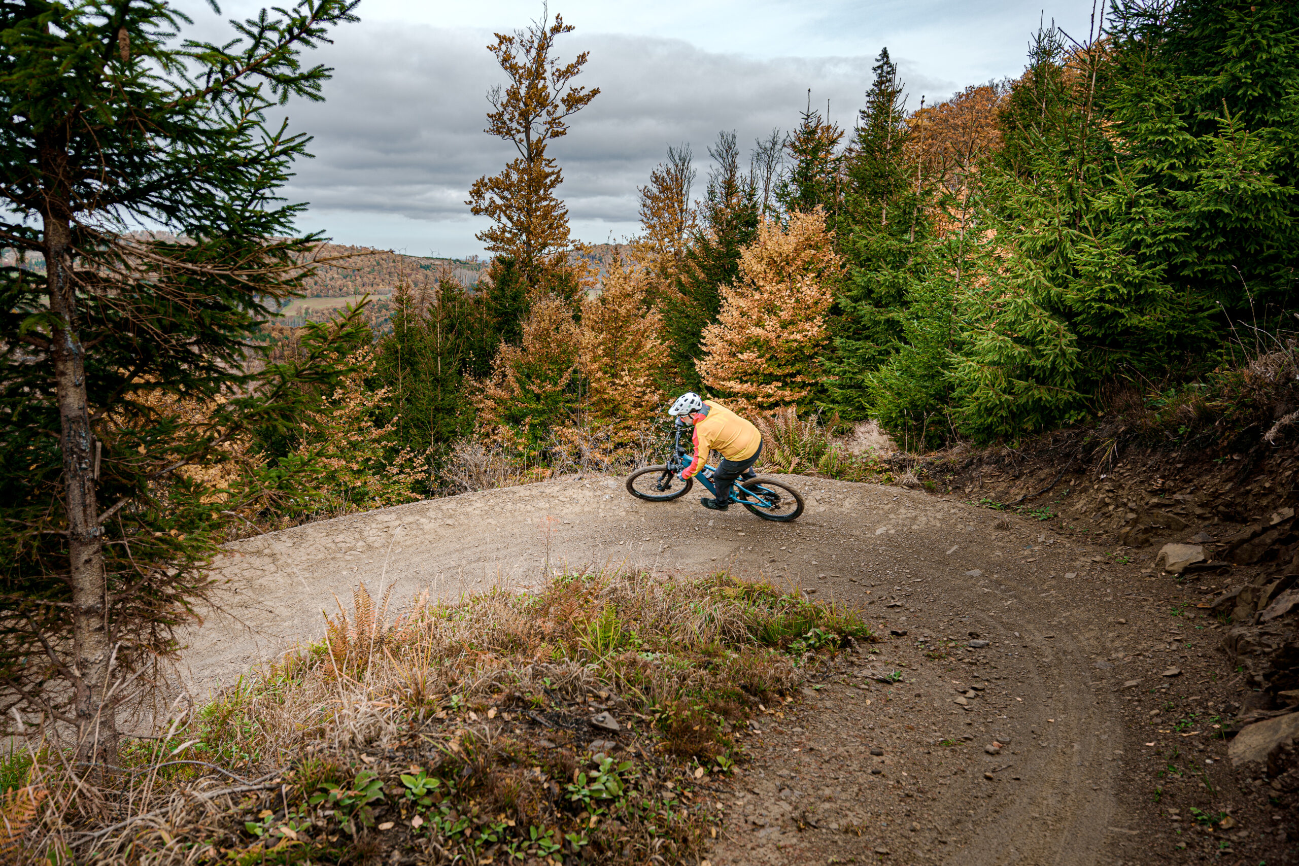 Ein Mountainbiker mit gelber Jacke und roten Ärmeln fährt allein mit einem blauen Mountainbike durch eine Kurve auf einem befestigten Weg. Der Trail ist von Bäumen umgeben und gut befahrbar. Auch trägt der Fahrer einen Helm. Die Szene wirkt dynamisch und vermittelt Bewegung und Konzentration. Ein Mountainbiker mit gelber Jacke und roten Ärmeln fährt allein mit einem blauen Mountainbike durch eine Kurve auf einem befestigten Weg. Der Trail ist von Bäumen umgeben und gut befahrbar. Auch trägt der Fahrer einen Helm. Die Szene wirkt dynamisch und vermittelt Bewegung und Konzentration.