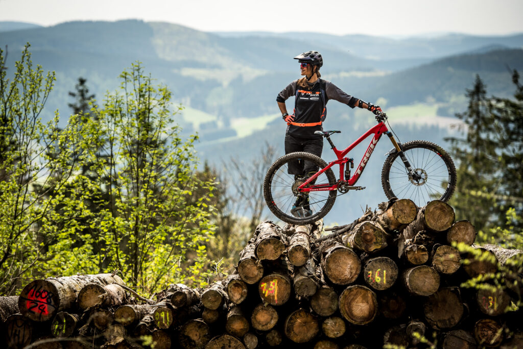 Eine Person mit Fahrrad steht auf gestapelten Baumstämmen im Außenbereich.
